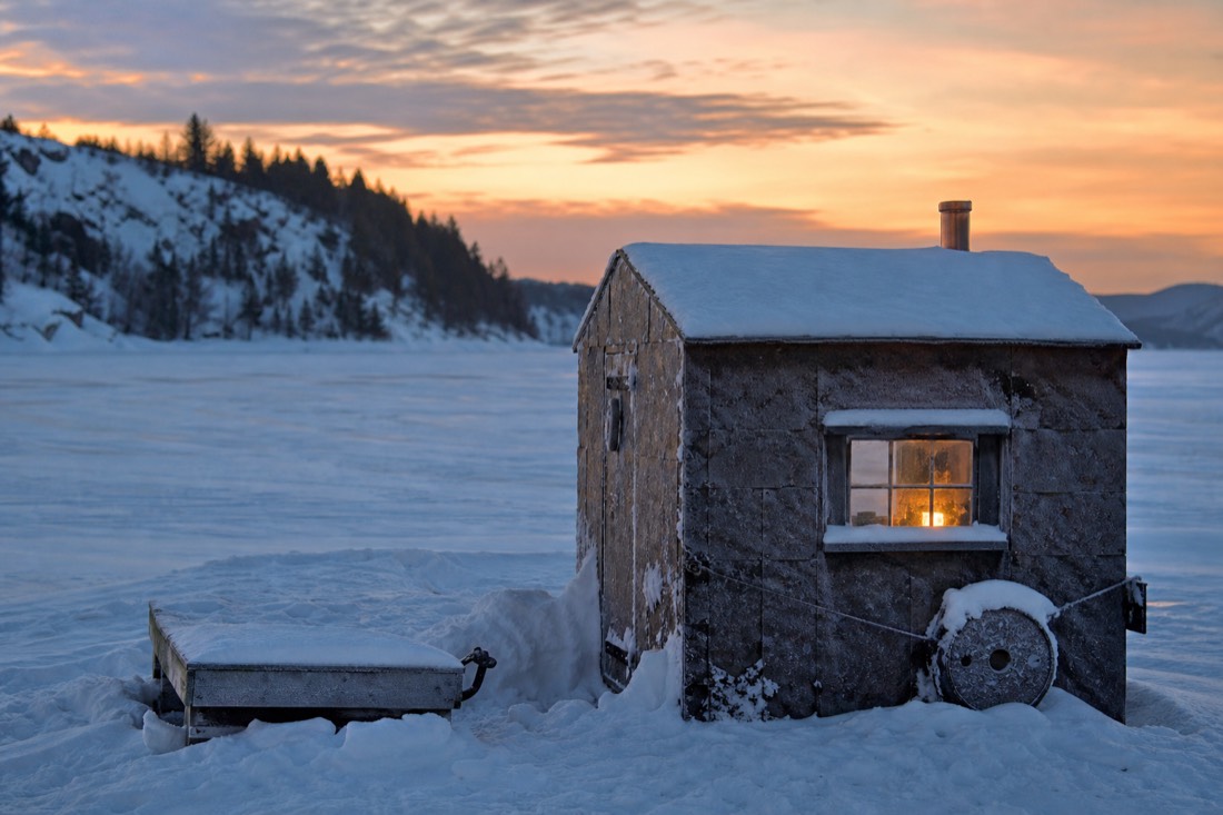 Snow-covered ice fishing cabin on a frozen Quebec river at dusk with warm light in the window