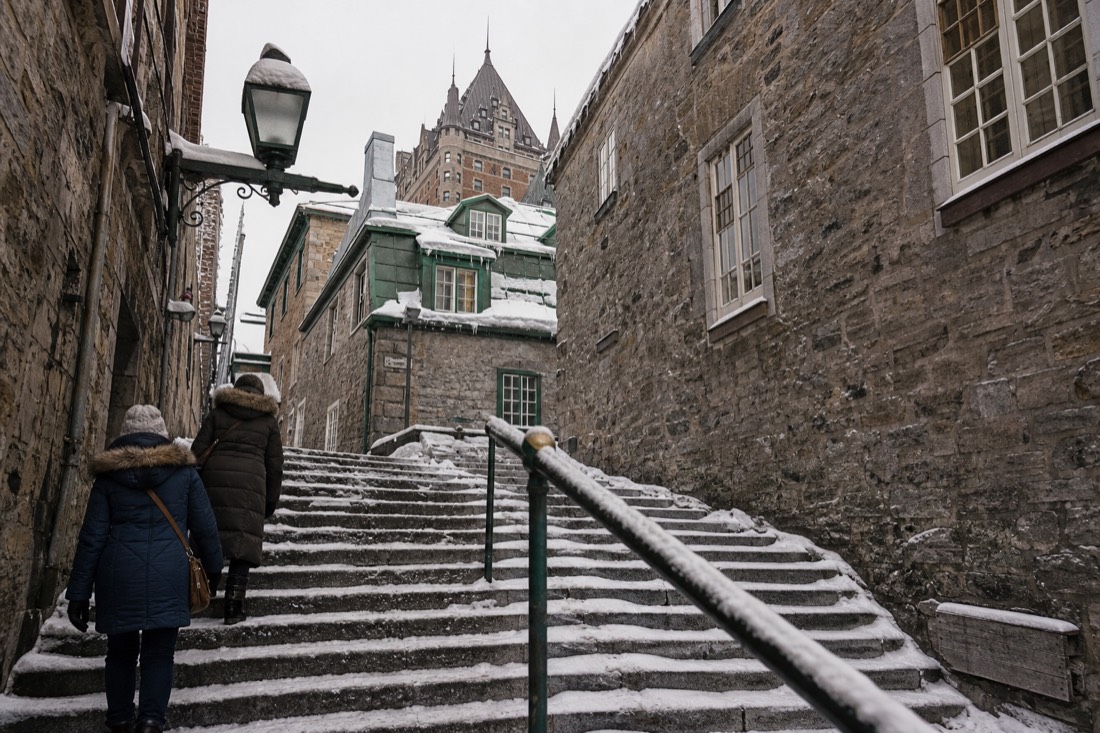 Winter visitors in long insulated coats and fur-trimmed hoods climbing a snowy staircase in Old Quebec